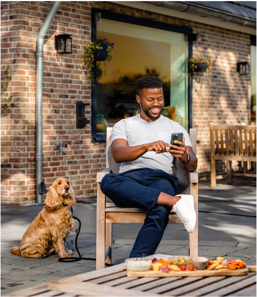 Man die op telefoon kijkt naar de opbrengsten van zijn zonnepanelen zittend in tuin met hond naast zich
