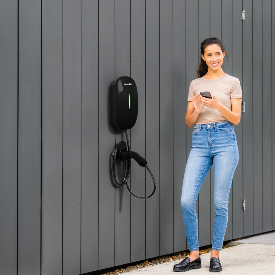 Vrouw op mobiel naast haar Charge 2 laadpaal, op de oprit, naast een Tesla Model 3